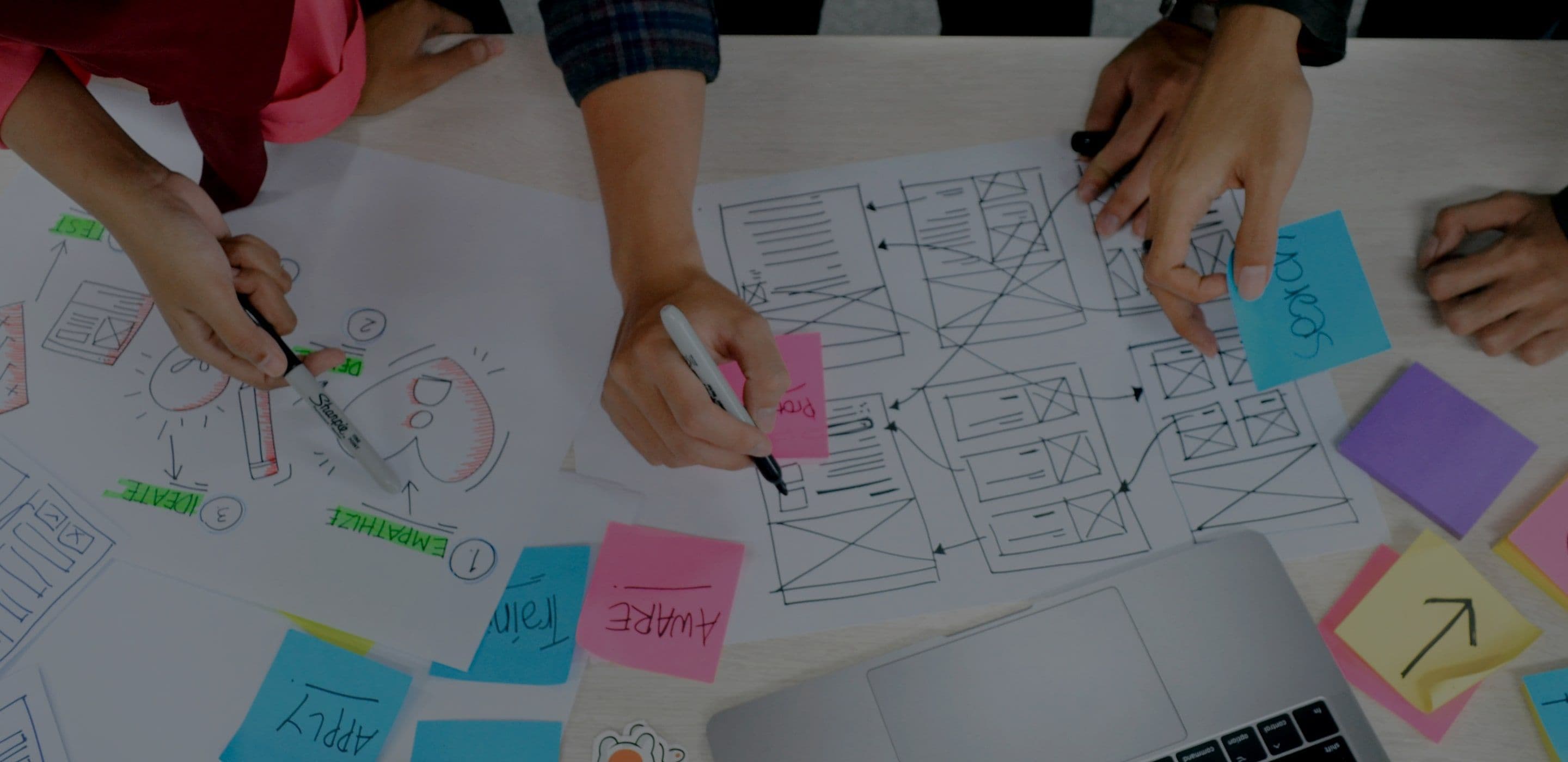 Hands sketching wireframes and user flows on paper beside a laptop