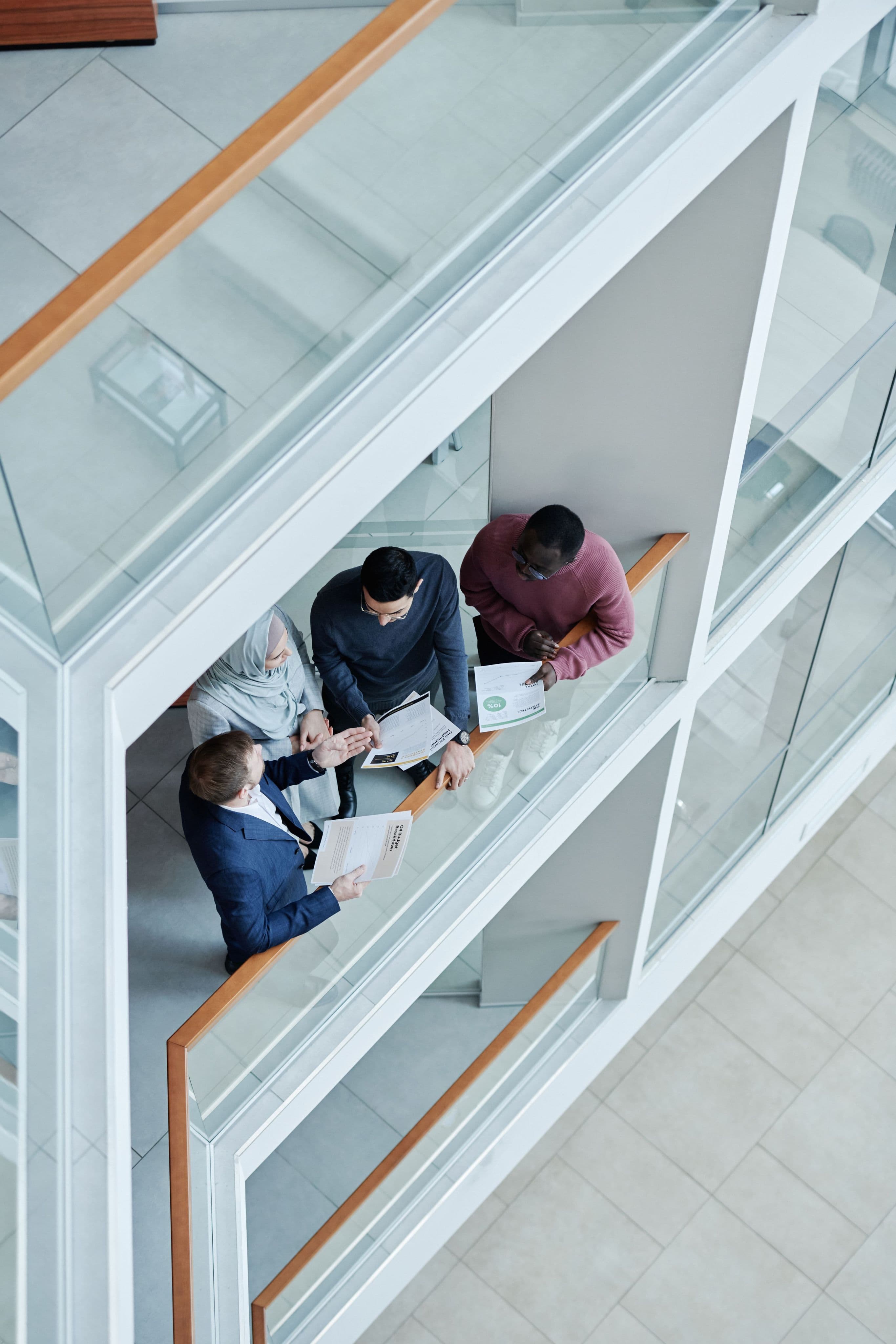 Four colleagues reviewing documents on a glass walkway, seen from above