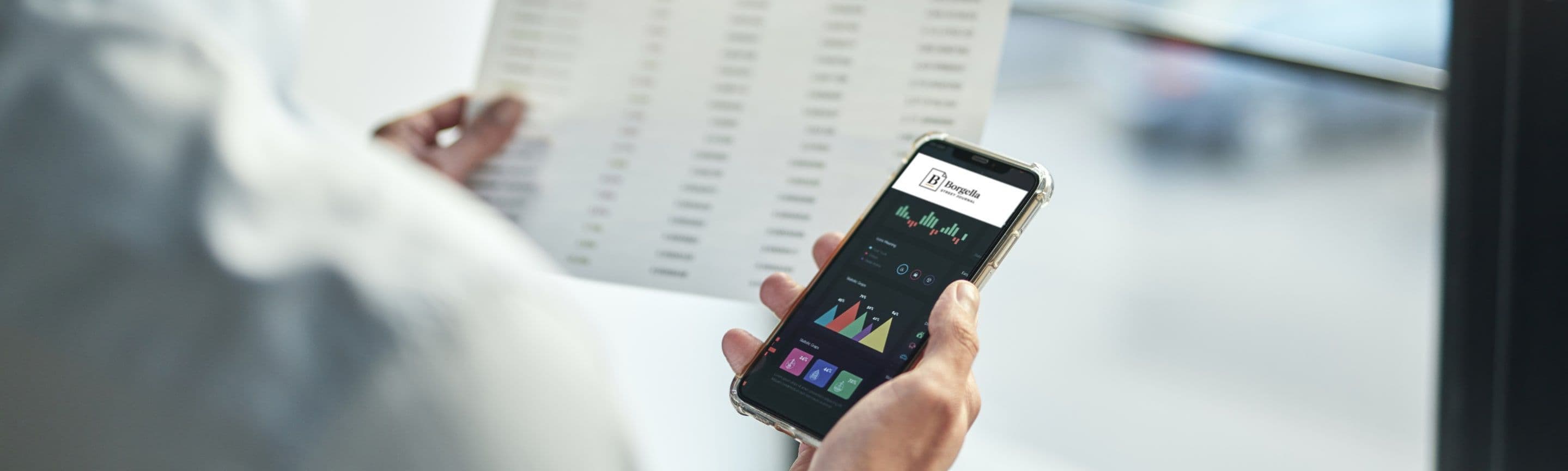 A hand holding a phone open on the Borgella app beside a printed report